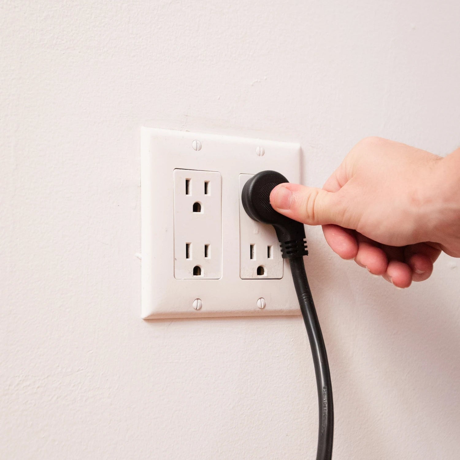 Hand plugging a black cord into a white electrical outlet on a light gray background